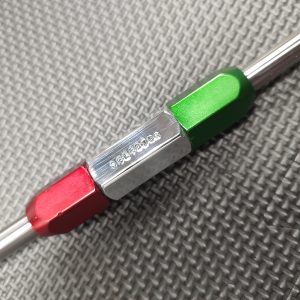 A close-up of the SOTAR Enhanced AR15 Extractor, a double-ended metal tool with red, silver, and green sections, lying on a textured dark gray surface.