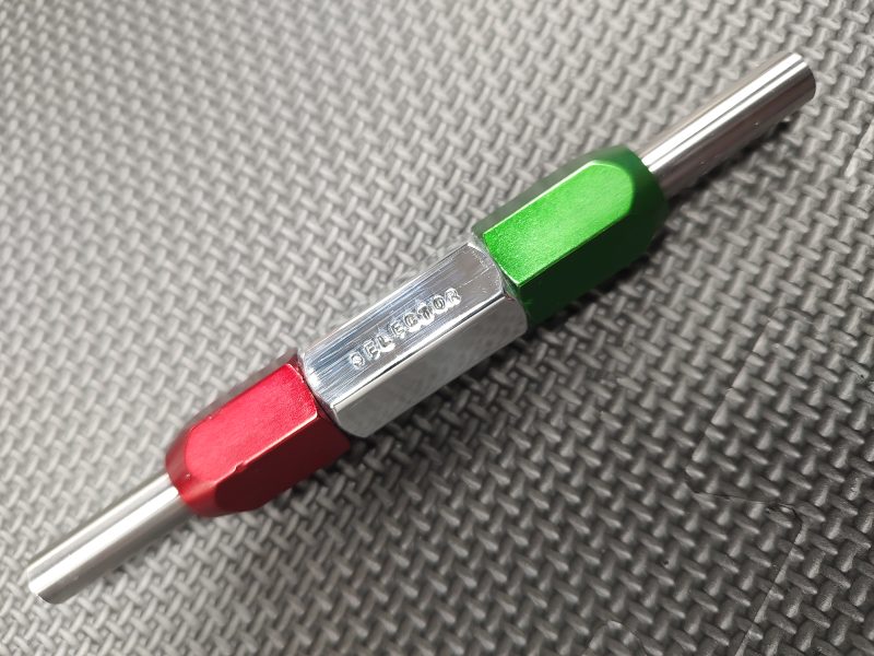 A close-up of the SOTAR Enhanced AR15 Extractor, a double-ended metal tool with red, silver, and green sections, lying on a textured dark gray surface.