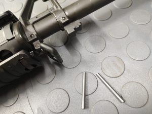 Close-up of the SOTAR Enhanced AR15 Extractor with two small metal pins on a textured surface beside the gun part.