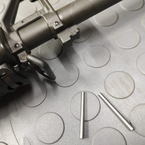 Close-up of the SOTAR Enhanced AR15 Extractor with two small metal pins on a textured surface beside the gun part.