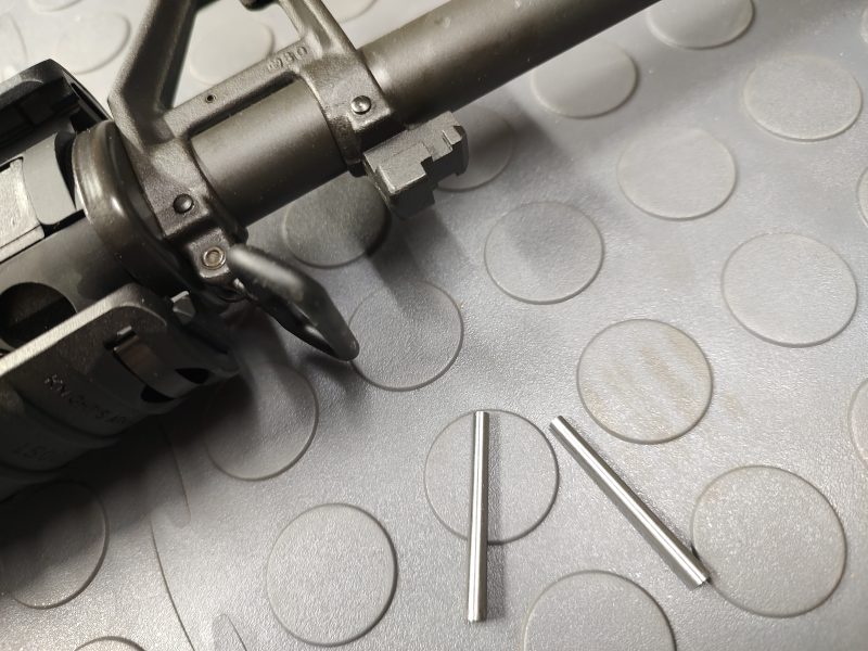Close-up of the SOTAR Enhanced AR15 Extractor with two small metal pins on a textured surface beside the gun part.