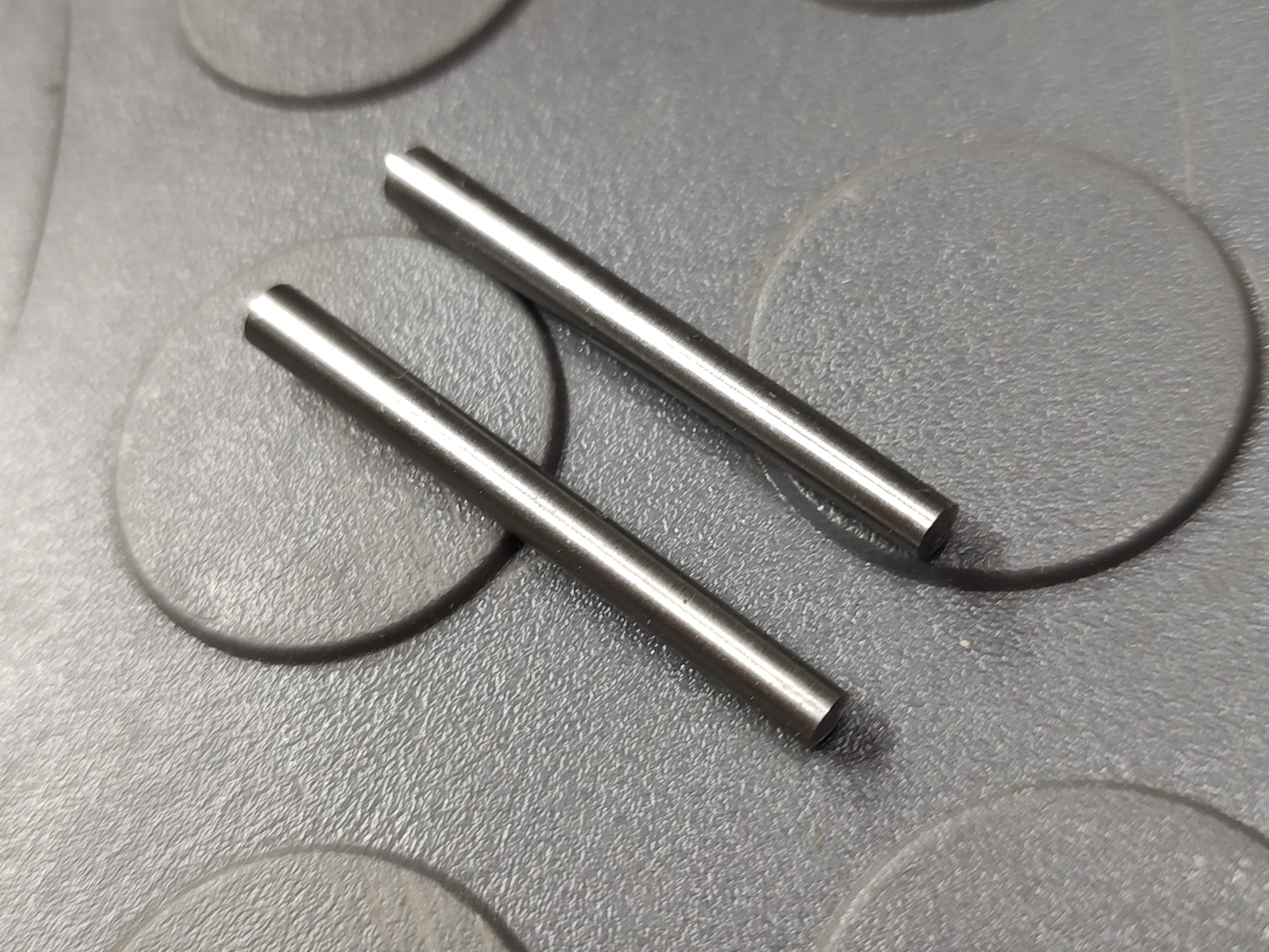 Two SOTAR Enhanced AR15 Extractors, both cylindrical metal components, are lying parallel to each other on a textured surface with a circular pattern.