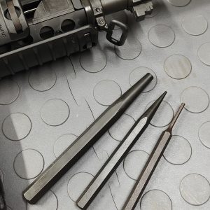 The SOTAR Enhanced AR15 Extractor is displayed beside three steel punches of varying sizes on a gray textured surface near the front end of a firearm.