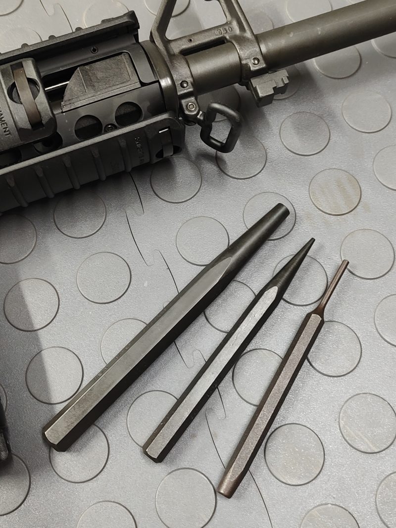 The SOTAR Enhanced AR15 Extractor is displayed beside three steel punches of varying sizes on a gray textured surface near the front end of a firearm.