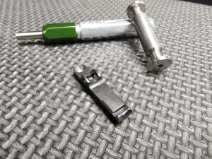 The SOTAR Enhanced AR15 Extractor, featuring a green handle, metal pin, and small metal component, is displayed on a textured gray surface.