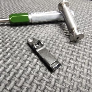 The SOTAR Enhanced AR15 Extractor, featuring a green handle, metal pin, and small metal component, is displayed on a textured gray surface.