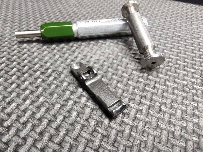 The SOTAR Enhanced AR15 Extractor, featuring a green handle, metal pin, and small metal component, is displayed on a textured gray surface.