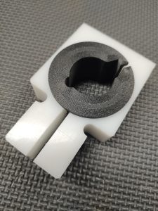 A close-up of the Magazine Button Install Tool, a white plastic rectangular part with a black circular insert and central cut-out, shown on a textured gray surface.