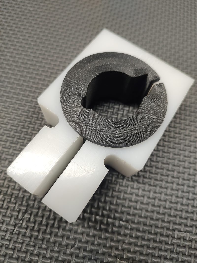 A close-up of the Magazine Button Install Tool, a white plastic rectangular part with a black circular insert and central cut-out, shown on a textured gray surface.