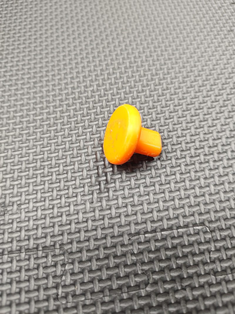 A Magazine Button Install Tool made of orange plastic rests on a textured black surface.