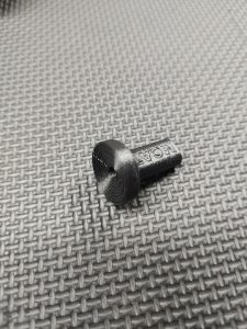 The Magazine Button Install Tool is a small black 3D-printed plastic item with a circular end and textured stem, shown on a woven gray surface.