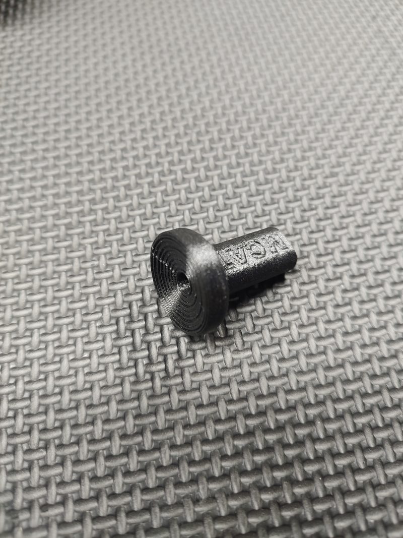 The Magazine Button Install Tool is a small black 3D-printed plastic item with a circular end and textured stem, shown on a woven gray surface.