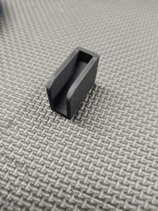 A compact, black, rectangular Magazine Button Install Tool made of plastic with an open slot on one side, shown on a textured gray surface.