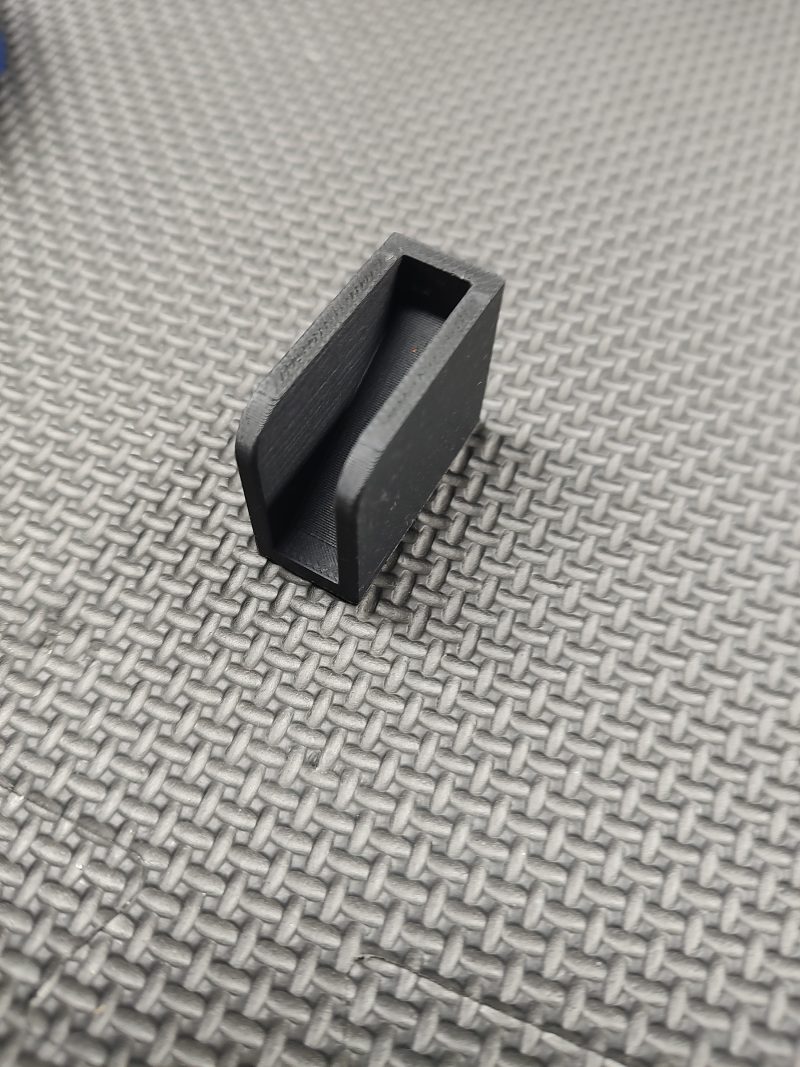 A compact, black, rectangular Magazine Button Install Tool made of plastic with an open slot on one side, shown on a textured gray surface.
