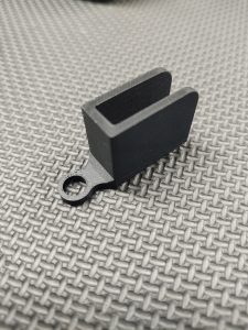 The Magazine Button Install Tool is a small, black, rectangular plastic piece with an open slot and a circular hole on one end, shown resting on a textured gray surface.