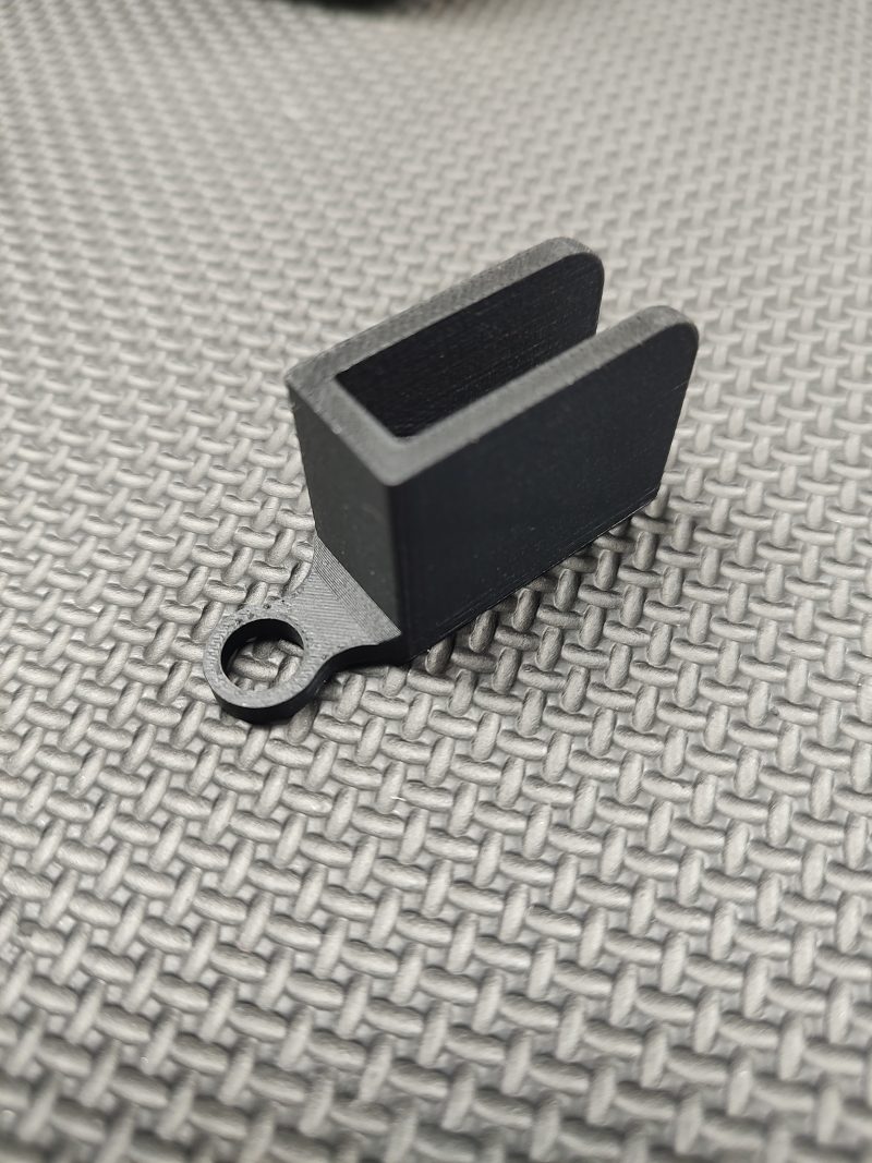The Magazine Button Install Tool is a small, black, rectangular plastic piece with an open slot and a circular hole on one end, shown resting on a textured gray surface.