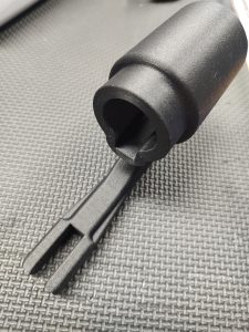 A close-up of the Magazine Button Install Tool, featuring a black plastic build with a cylindrical top and pronged end, shown resting on a textured dark surface.
