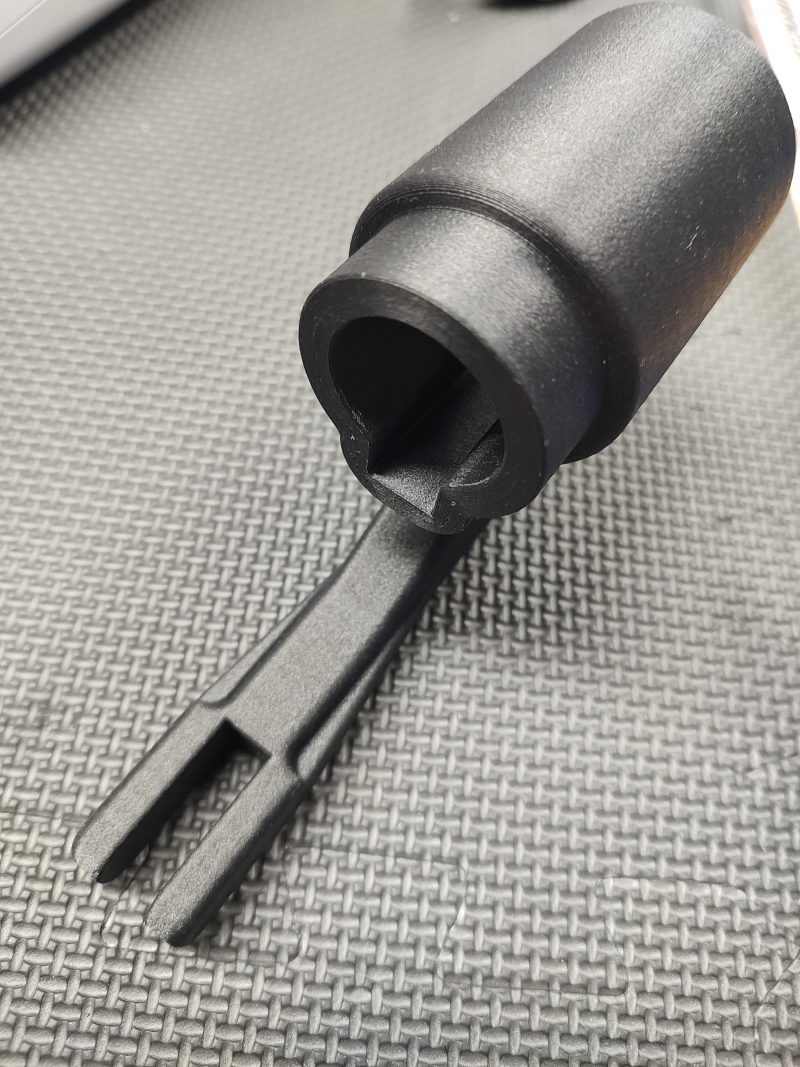 A close-up of the Magazine Button Install Tool, featuring a black plastic build with a cylindrical top and pronged end, shown resting on a textured dark surface.