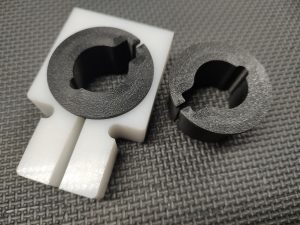 The Magazine Button Install Tool consists of a black circular piece and a black ring set into a white rectangular base with a circular cutout on a textured surface.