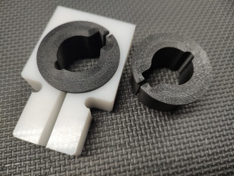 The Magazine Button Install Tool consists of a black circular piece and a black ring set into a white rectangular base with a circular cutout on a textured surface.