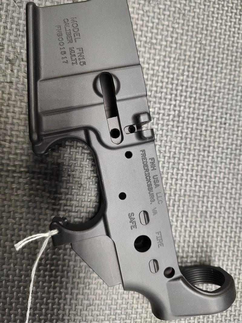 A close-up of the Stripped FN15 Lower Receiver - Fully Inspected and Gauged, a black metal firearm component, resting on a textured gray surface.