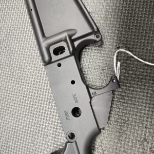 The Stripped FN15 Lower Receiver - Fully Inspected and Gauged is shown on a textured gray surface with a tag attached to its trigger guard.