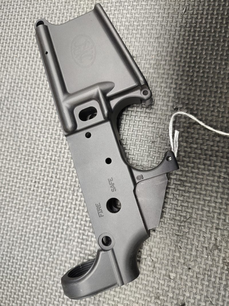 The Stripped FN15 Lower Receiver - Fully Inspected and Gauged is shown on a textured gray surface with a tag attached to its trigger guard.