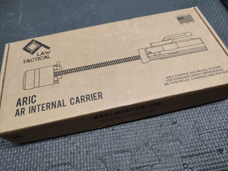 A cardboard box labeled "LAW Tactical ARIC - C Model" with a line drawing of the product and website URL, placed on a textured dark surface.