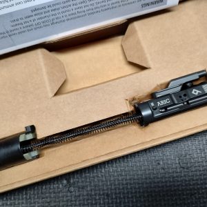 A close-up of the LAW Tactical ARIC - C Model bolt carrier group with a recoil spring, inside a cardboard box and partially covered by packaging material.