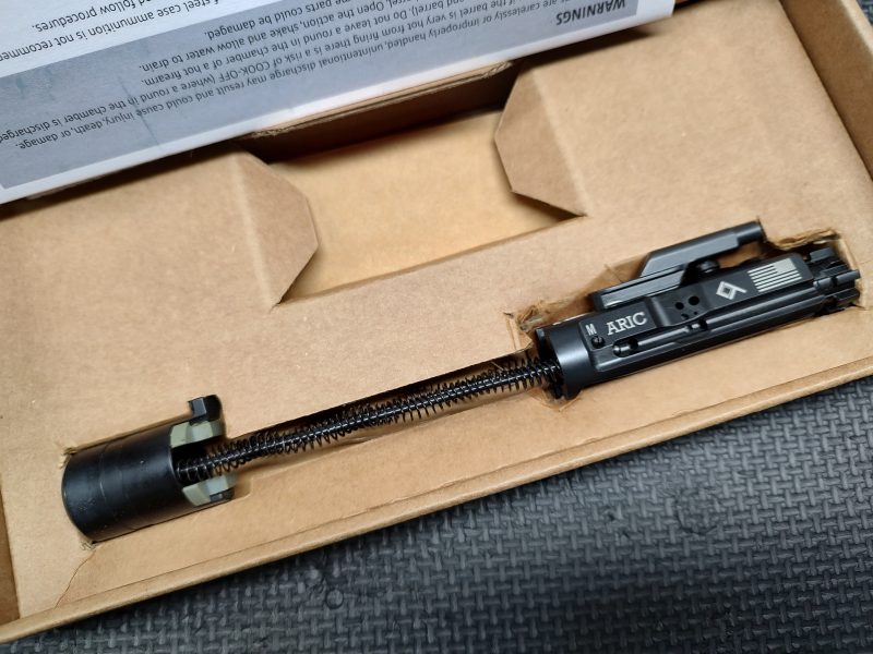 A close-up of the LAW Tactical ARIC - C Model bolt carrier group with a recoil spring, inside a cardboard box and partially covered by packaging material.
