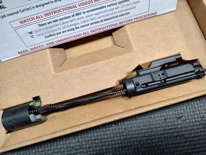 The LAW Tactical ARIC - C Model, featuring an attached spring assembly, is placed inside a cardboard box with partially visible instructions in the background.