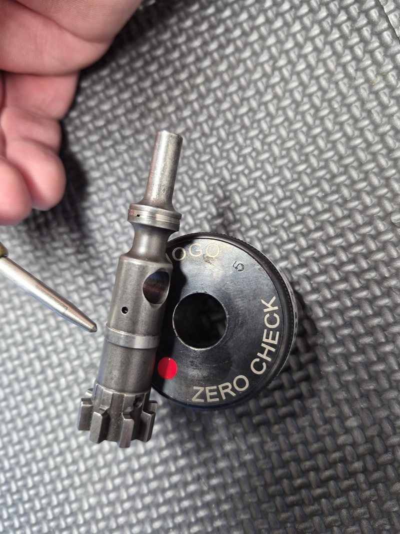 The SOTAR AR15 NEW Bolt Support Ring NOGO Gauge sits beside a circular "ZERO CHECK" tool on a textured black surface, with part of a hand and metallic pen also in view.
