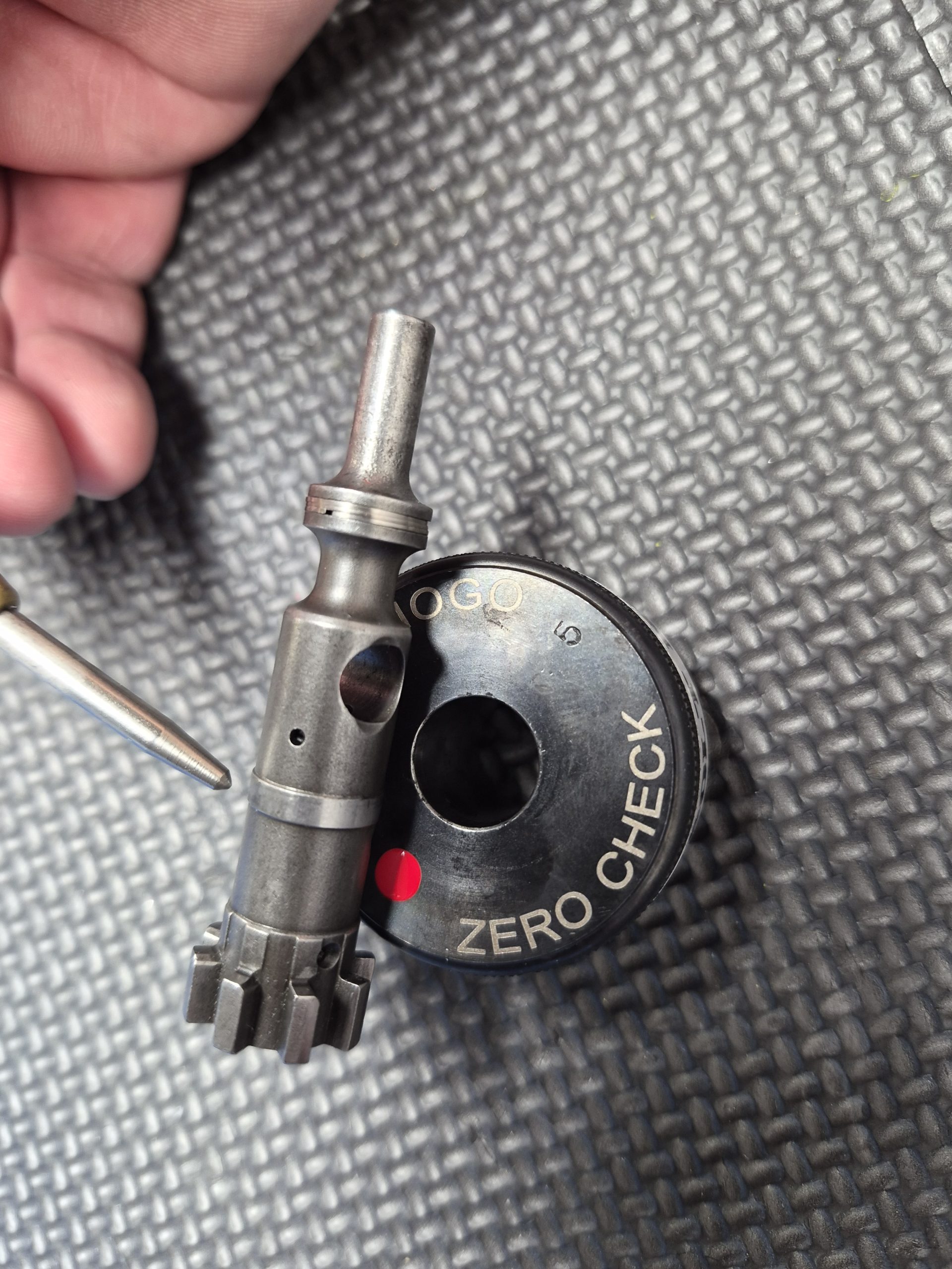 The SOTAR AR15 NEW Bolt Support Ring NOGO Gauge sits beside a circular "ZERO CHECK" tool on a textured black surface, with part of a hand and metallic pen also in view.