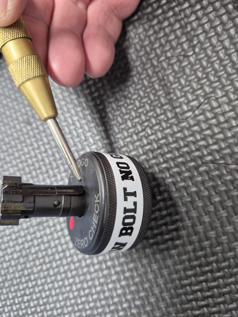 A hand holds a brass punch tool pressing the SOTAR AR15 NEW Bolt Support Ring NOGO Gauge, labeled "BOLT NO GO," on a textured black surface.