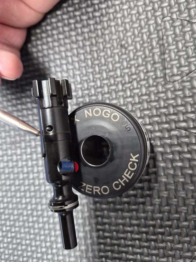 A close-up of the SOTAR AR15 NEW Bolt Support Ring NOGO Gauge shows its black mechanical part on the round gauge labeled "NOGO" and "ZERO CHECK," with a hand and textured surface in the background.