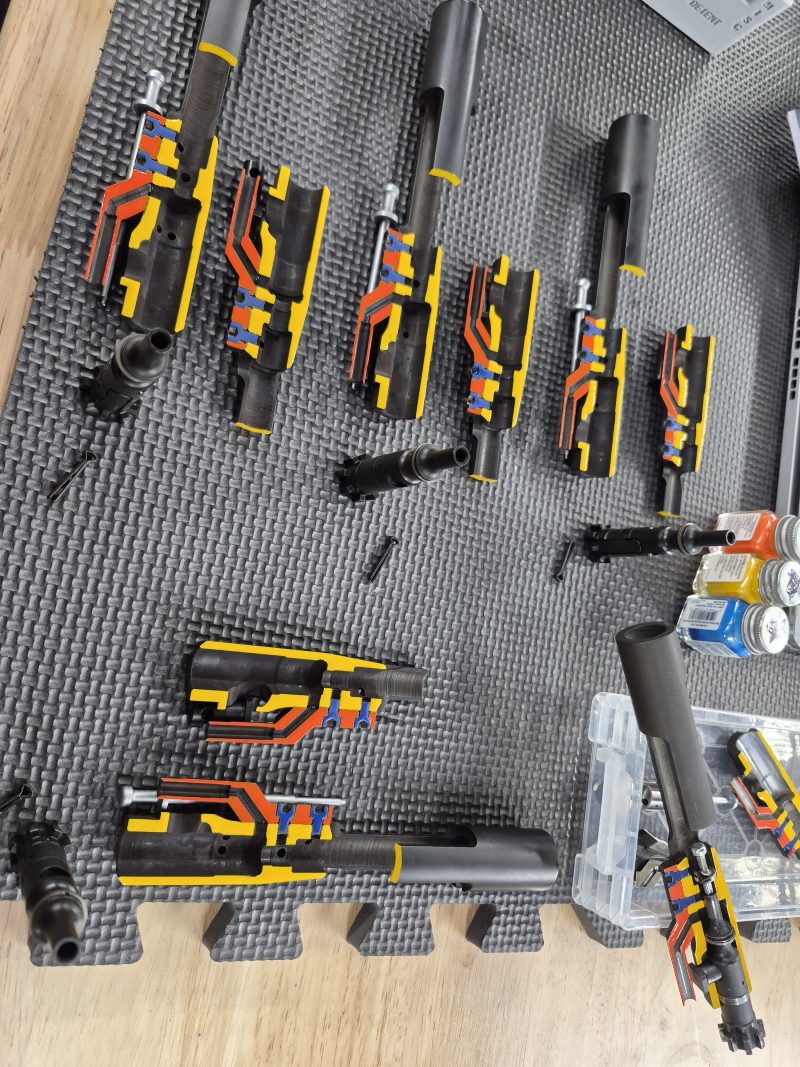 An EDM CUT - CUTAWAY M16 Bolt Carrier Group with attached components is arranged on a textured mat, alongside small tools and containers on a wooden surface.
