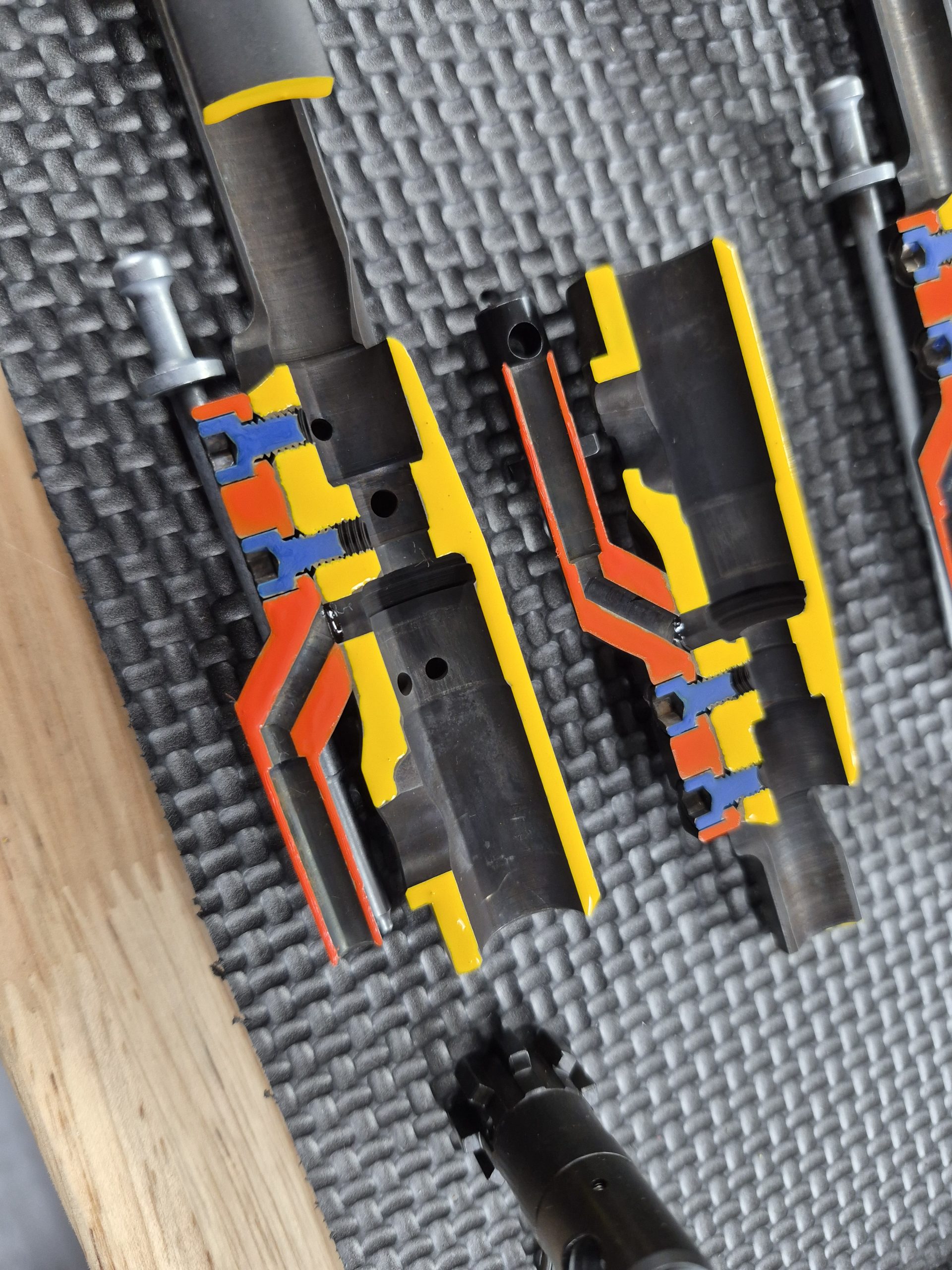 Two EDM CUT - CUTAWAY M16 Bolt Carrier Groups with colored cross-sections highlighting their internal pathways and chambers are placed on a textured black mat.