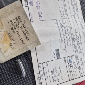 Displayed on a textured black surface is the USGI M1911 Headspace Gauge - RARE/COLLECTORS ITEM, featuring a metal gage with a brown label, a white return gage card, and a certification envelope with handwritten notes.