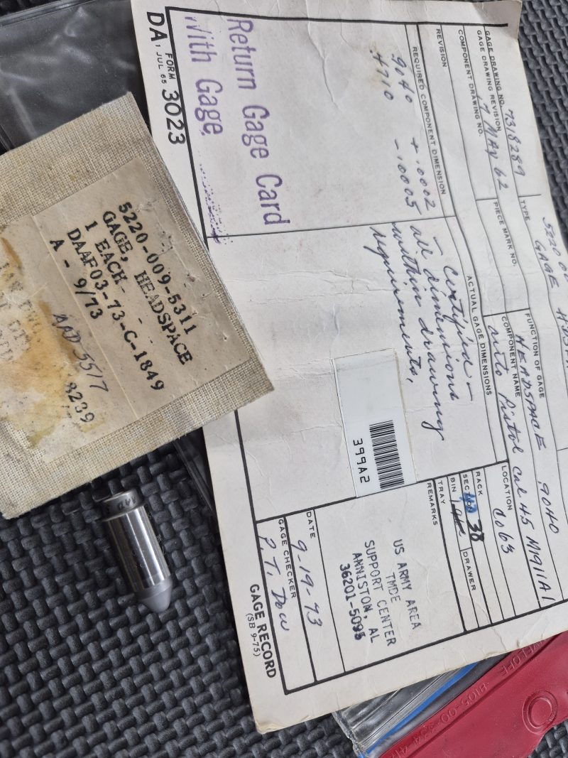 Displayed on a textured black surface is the USGI M1911 Headspace Gauge - RARE/COLLECTORS ITEM, featuring a metal gage with a brown label, a white return gage card, and a certification envelope with handwritten notes.
