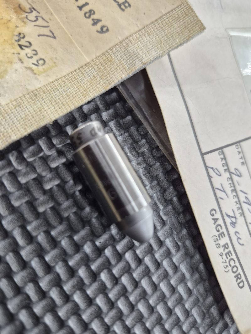 A USGI M1911 Headspace Gauge—a rare collector’s item—rests on a textured black surface beside papers with handwritten and typed notes.