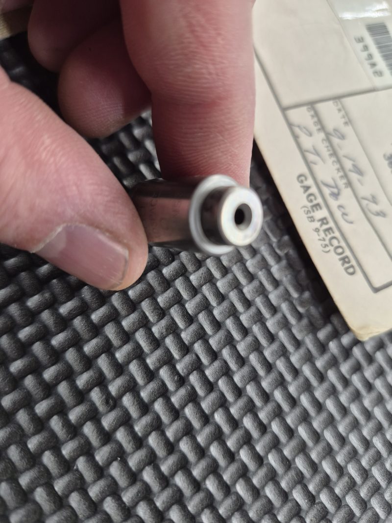 A hand holds a USGI M1911 Headspace Gauge—an uncommon collector’s item—featuring a metal cylindrical form with a small central hole, positioned above a textured black surface and near a paper labeled “GAGE RECORD.”.