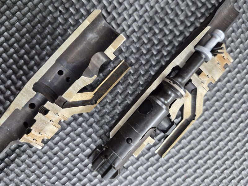 Two EDM CUT - CUTAWAY M16 Bolt Carrier Groups are displayed on a textured black surface, revealing their internal components and mechanisms.