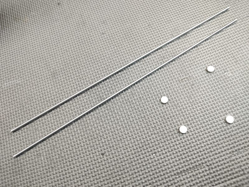 Two Muzzle Device Alignment Rods with Rare Earth Retention Magnets are placed parallel on a textured surface with five small, round magnets arranged to the right of the rods.