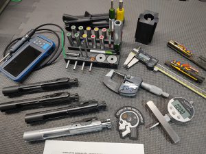 A collection of tools and measuring instruments including calipers, micrometer, SOTAR AR15 BCG 3 Bore & Gas System Gauge Set with Imported Handle - BACKORDER, screwdrivers, and various gun parts arranged on a textured black surface.