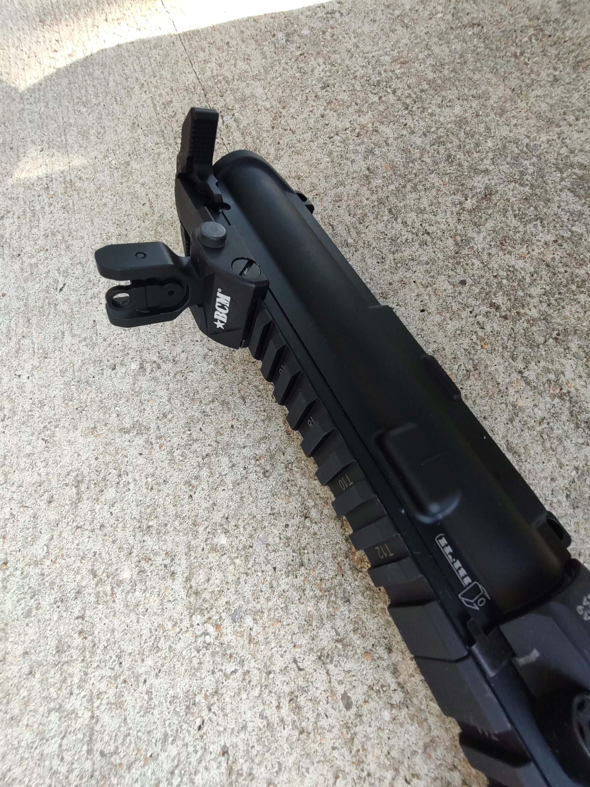 A close-up view of a BCM® COMPLETE 16" CHF Mid Length Complete Upper Receiver Group w/ MCMR-9 Handguard - New Old Stock - Minor Handling Marks attached to a rail mount on a concrete surface, with iron sights flipped up.