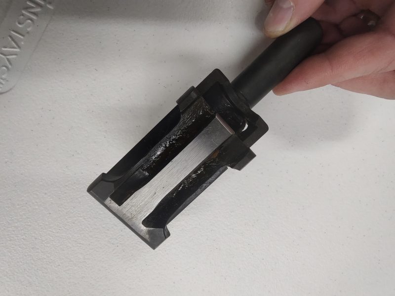 A hand holding the HK33 Bolt Carrier, a black metal tool with a rectangular shape and multiple grooves, against a white background.