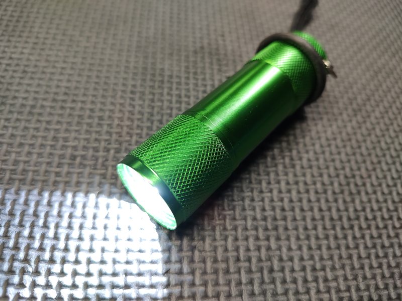 A green AR Upper Receiver Illumination Tool is turned on, emitting light and lying on a textured grey surface.