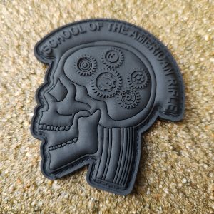 SOTAR PVC Patch - MK3 - Subdued (Black on Black) features a black patch displaying a profile of a skull with gears inside the brain area, surrounded by the text "School of the American Rifle" at the top. The background is textured sand.