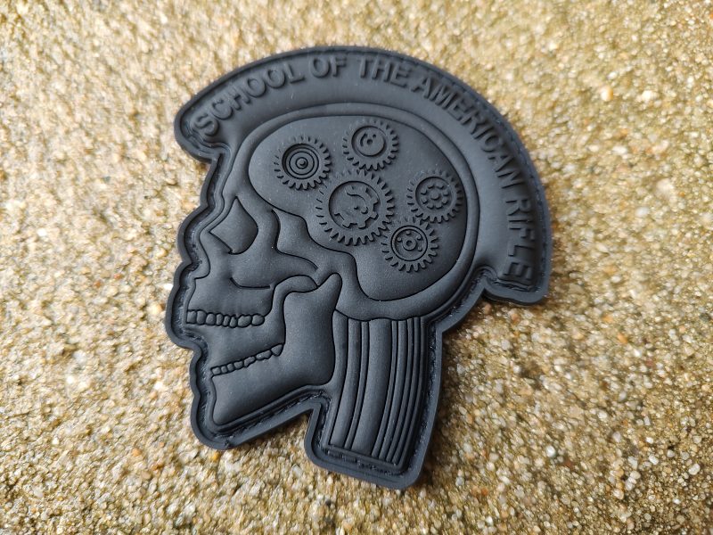 SOTAR PVC Patch - MK3 - Subdued (Black on Black) features a black patch displaying a profile of a skull with gears inside the brain area, surrounded by the text "School of the American Rifle" at the top. The background is textured sand.