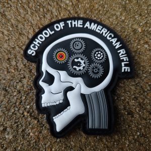 A "SOTAR PVC Patch - MK2 White," featuring a side profile of a skeleton's head filled with gears and labeled "School of the American Rifle," placed on a rough textured surface.